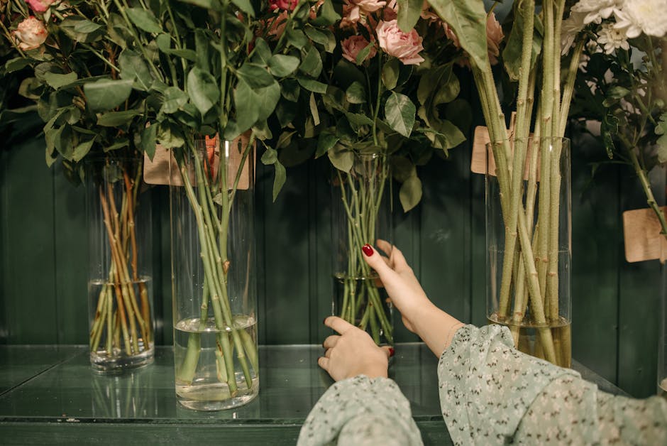 A person arranging a bouquet of mixed flowers, including pink roses and lush green foliage, in a tall, clear glass vase. The arrangement is placed on a green surface against a dark green wall, with se