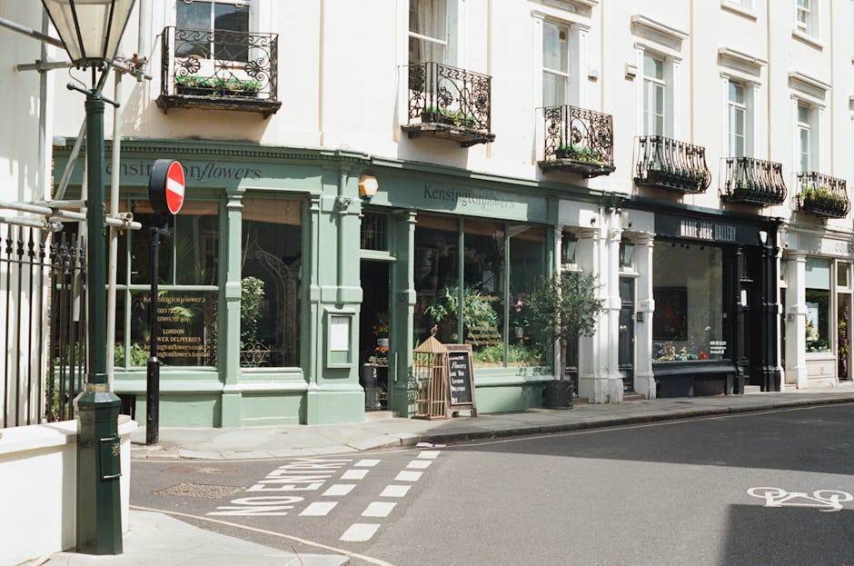 A storefront of a flower shop on Kensington High Street featuring a pastel green facade with large glass windows displaying floral arrangements inside. The shop is adorned with small balcony flower bo