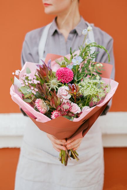 A person dressed in a grey shirt and white apron holds a bouquet of fresh flowers wrapped in soft pink tissue paper and brown craft paper, standing against an orange background. The bouquet features a