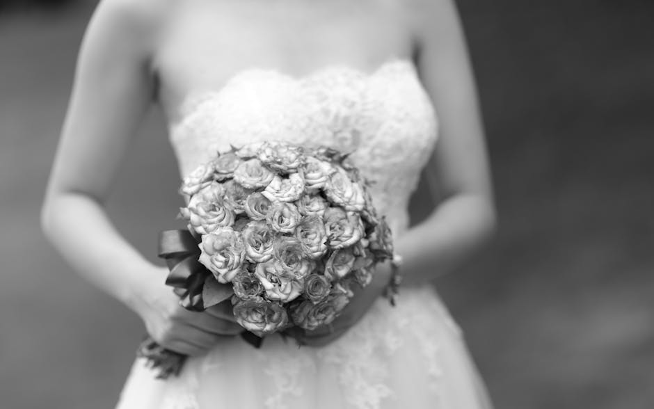 A bride dressed in a strapless white lace wedding gown holds a bouquet of tightly clustered roses, featuring soft petals and lush green stems, wrapped in a satin ribbon. The bouquet is positioned at w