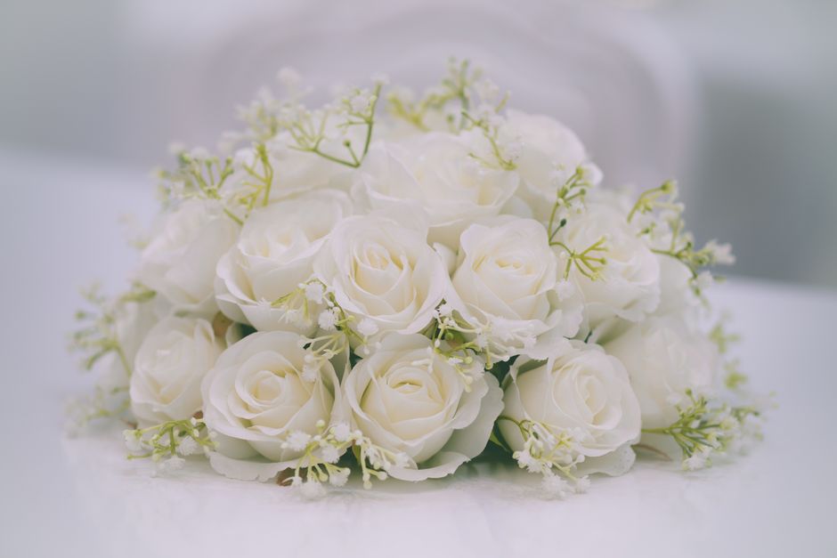 A bouquet composed of creamy white roses with delicate, softly curled petals arranged in a rounded, compact style. Interspersed among the roses are small sprigs of white baby's breath with tiny, star-