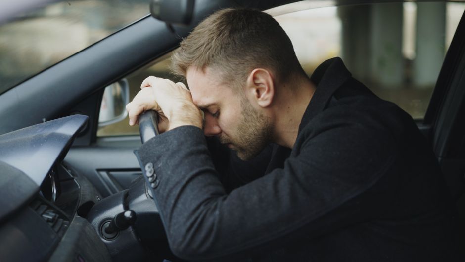 A man with light brown hair and a beard, dressed in a dark coat, is seated inside a car. He appears distressed, with his forehead resting on the steering wheel and his hands clasped over his head. The