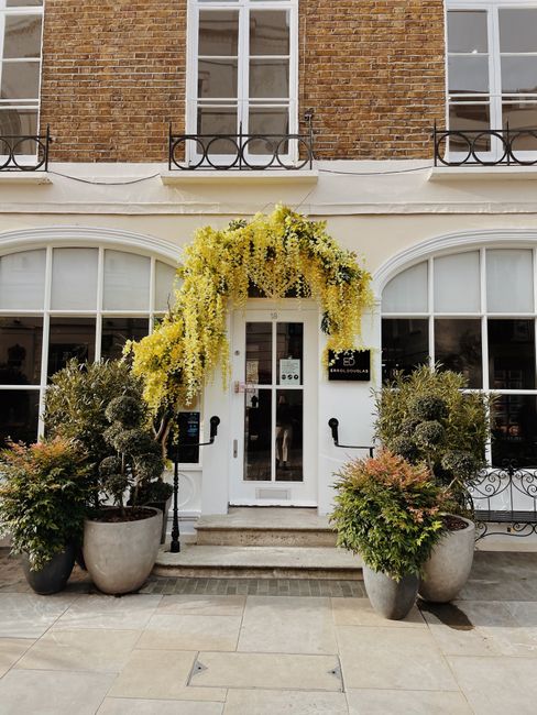 The entrance of a building decorated with a lush floral arrangement featuring cascading yellow flowers, possibly wisteria or similar blooms, draping over the white doorway. Flanking the entrance are t