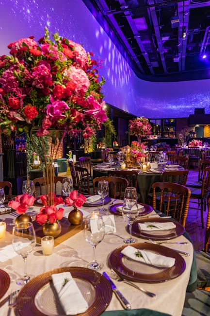 A floral arrangement featuring a large, vibrant bouquet of pink and red flowers, including roses and hydrangeas, displayed atop a tall, slender gold stand. The bouquet's petals are fresh and slightly