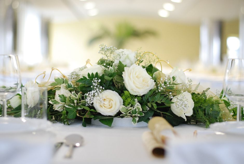 A floral arrangement featuring a bouquet of white roses, delicate baby's breath, and green foliage, arranged in a low, elongated style on a table. The bouquet is surrounded by glassware and table sett