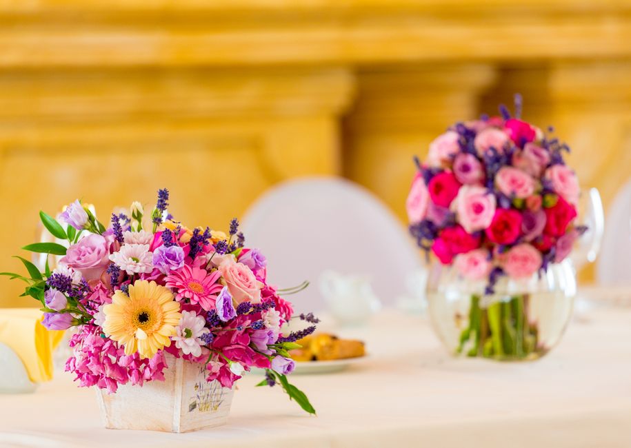 A floral arrangement featuring a vibrant mix of pink, purple, and yellow flowers, including gerbera daisies, roses, and chrysanthemums, arranged in a rustic white wooden container. The bouquet display