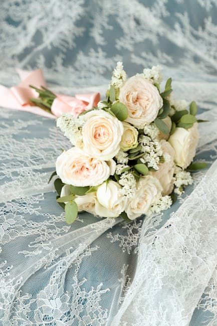 A delicate bouquet of fresh flowers resting on light blue lace fabric, featuring soft pink and white roses, small white filler flowers, and green foliage. The bouquet is wrapped in pale pink paper wit