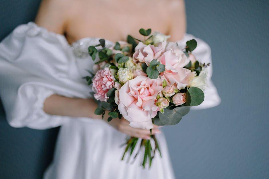 A person dressed in a white, off-the-shoulder garment holds a delicate floral bouquet featuring large, soft pink roses, white lisianthus, and light pink carnations, accented with fresh, rounded green 