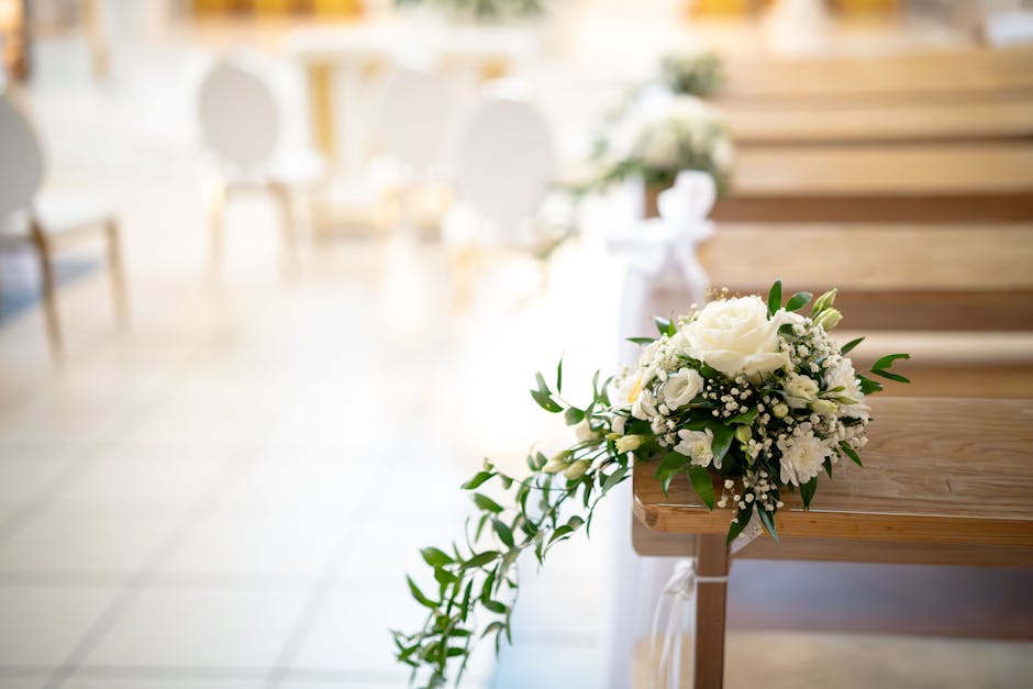 A white floral bouquet featuring roses, baby's breath, and greenery resting on the edge of a wooden pew, with soft natural light illuminating the arrangement in an indoor setting. The bouquet displays